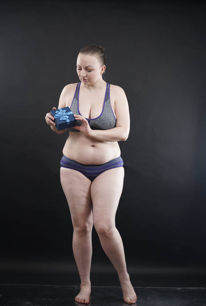 Hermosa mujer de tamaño grande que usa ropa interior deportiva sostiene la caja con suscripción de regalo a un gimnasio en fondo sólido estudio negro
 - Foto, imagen