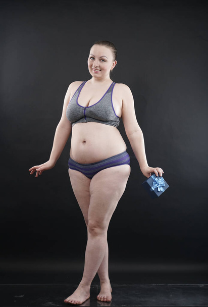 Beautiful plus size woman wearing sport underweaer holds box with gift subscription to a fitness on black studio solid background - Valokuva, kuva