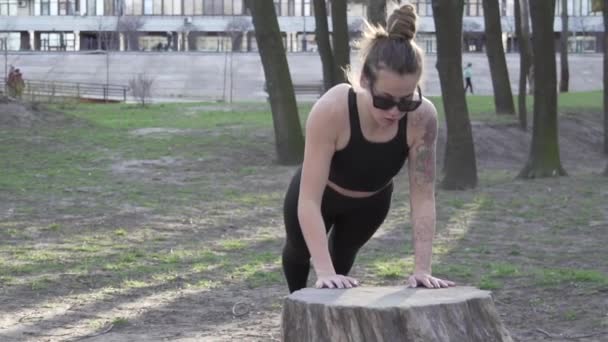 Empurre para cima ou pressione o exercício pela jovem mulher. Jovem fazendo flexões nas madeiras. Atleta fêmea fazendo exercícios para músculos do braço push-ups de uma árvore derrubada em um parque - Filmagem, Vídeo