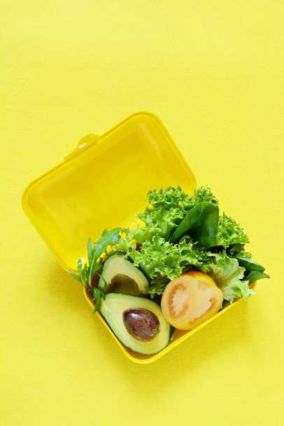 Lunchbox with a snack from lettuce, tomato, avacado on a yellow background. Healthy eating concept. Go vegan. Tasty vegetarian food in plastic box. - Photo, Image