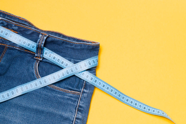 blue measuring tape is threaded into jeans instead of a belt, blue jeans on a yellow background, copy space, weight loss and weight control concept, measuring body volume - Foto, afbeelding