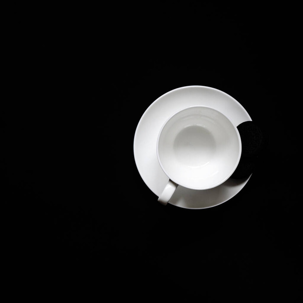 A white cup of coffee with black biscuits on a plate stands on a black background. Top view - 写真・画像