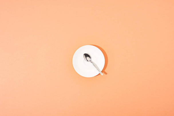 top view of metal spoon on white round plate on orange background - Foto, imagen