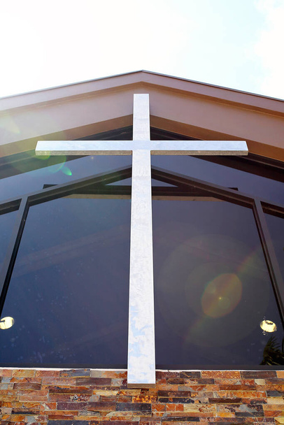 Ένα μεγάλο ασημένιο μέταλλο Σταυρός του Ιησού κρέμεται στο καμπαναριό πάνω από την μπροστινή είσοδο ενός θρησκευτικού κτιρίου Χριστιανική Εκκλησία. - Φωτογραφία, εικόνα