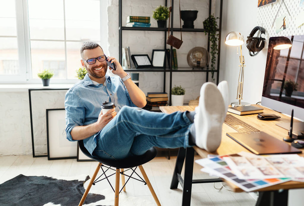 Vista lateral de alegre jovem do sexo masculino em uso casual e óculos falando ao telefone enquanto sentado com café takeaway no local de trabalho com grande monito
 - Foto, Imagem