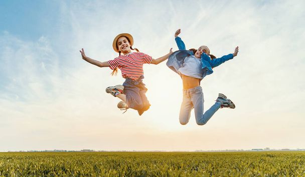 Снизу веселые маленькие сестры в повседневной одежде веселятся и прыгают с поднятыми руками на зеленом поле в летний вечер
 - Фото, изображение