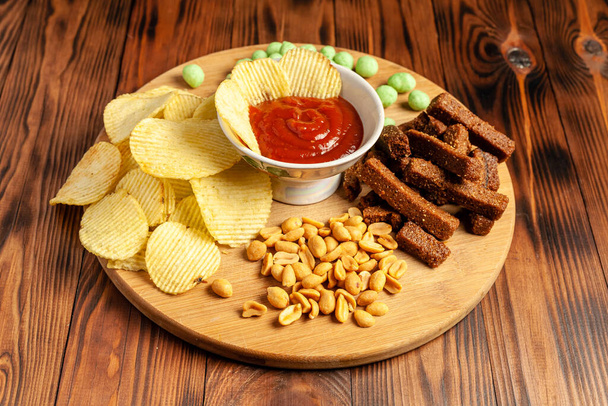 Verschiedene trockene Snacks auf einem Holzbrett. Chips, Erdnüsse, Croutons, Wasabi. - Foto, Bild
