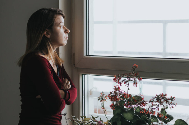 Νεαρή ανήσυχη γυναίκα κοιτάζει από το παράθυρο - Φωτογραφία, εικόνα