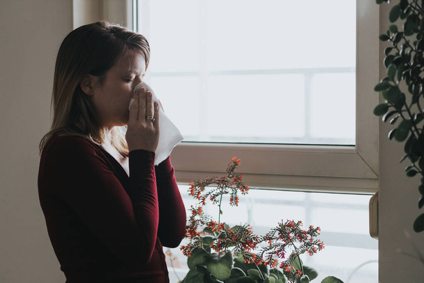 Mladá nemocná žena fouká nos do kapesníku u okna  - Fotografie, Obrázek