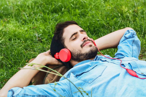 Portret młodego mężczyzny leżącego na trawie i słuchającego muzyki ze słuchawkami na zewnątrz.   - Zdjęcie, obraz