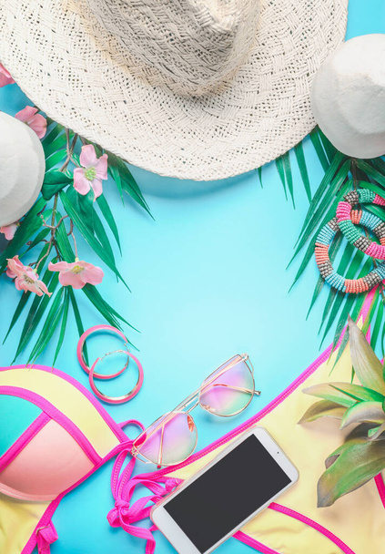 Top view of female beach accessories  background with tropical leaves and flowers , sunglasses and straw hat, bikini and smartphone on sunny blue. Summer holiday concept - Foto, Imagem