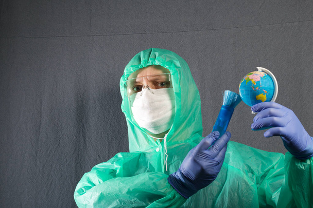 A man in a protective gauze mask, glasses and a raincoat with a hood. Holds a globe in his hand and brushes it with a brush. Hands in rubber gloves. Symbol of the global epidemic. It is on a gray background. - Foto, Bild