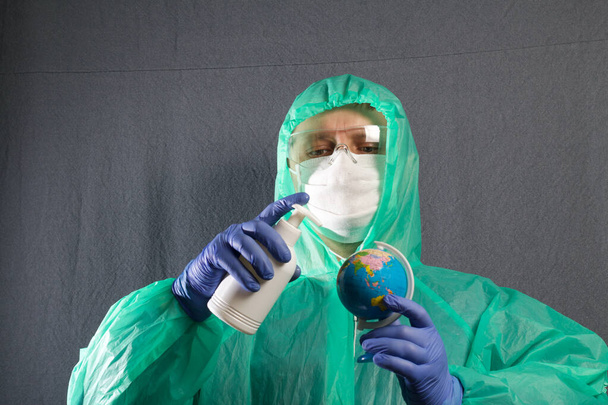 A man in a protective gauze mask, glasses and a raincoat with a hood. He holds the globe in his hand and processes it from the atomizer. Hands in rubber gloves. Symbol of the global epidemic. It is on a gray background. - Fotoğraf, Görsel