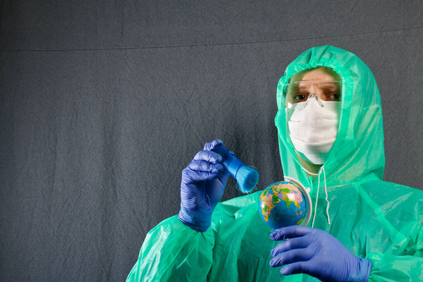A man in a protective gauze mask, glasses and a raincoat with a hood. Holds a globe in his hand and brushes it with a brush. Hands in rubber gloves. Symbol of the global epidemic. It is on a gray background. - Fotografie, Obrázek