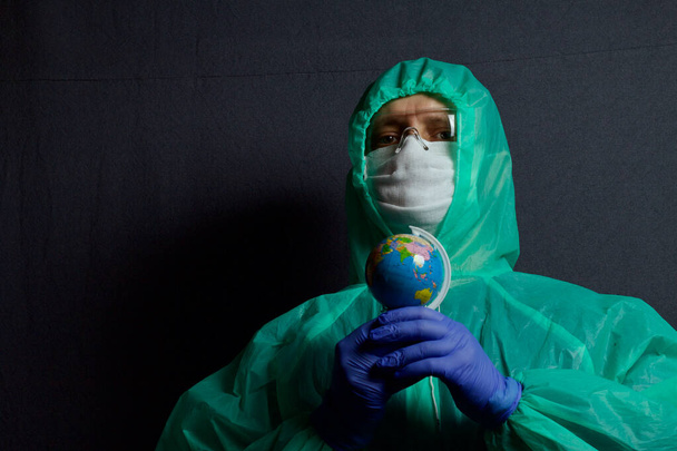 A man in a protective gauze mask, glasses and a raincoat with a hood. Holding a globe in his hands. Hands in rubber gloves. Symbol of the global epidemic. It is on a gray background. - 写真・画像