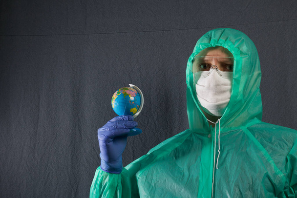 A man in a protective gauze mask, glasses and a raincoat with a hood. Holding a globe in his hand. Hands in rubber gloves. Symbol of the global epidemic. It is on a gray background. - Photo, Image