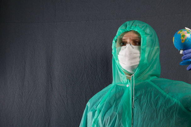 A man in a protective gauze mask, glasses and a raincoat with a hood. Holding a globe in his hand. Hands in rubber gloves. Symbol of the global epidemic. It is on a gray background. - Фото, зображення