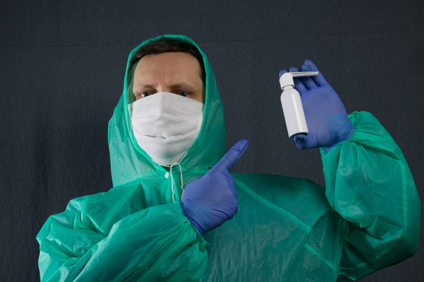 A man in a protective gauze mask and a raincoat with a hood. Holds a throat spray in his left hand, points to it with his right hand. Hands in rubber gloves. Simple protection against viral infection. It is on a gray background. - Photo, image