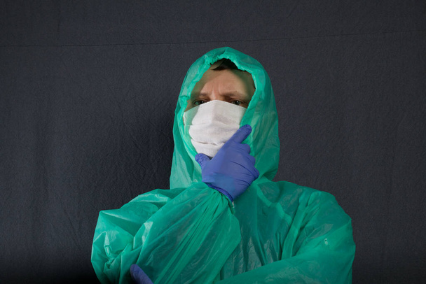 A man in a protective gauze mask and a raincoat with a hood. In rubber gloves. Pensively took up his chin. Simple protection against viral infection. It is on a gray background. - Foto, immagini