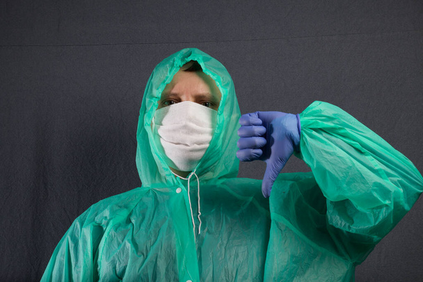 A man in a protective gauze mask and a raincoat with a hood. In rubber gloves. The thumb of the left hand is down. Simple protection against viral infection. It is on a gray background. - Foto, Imagem