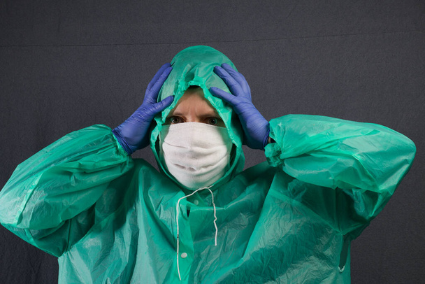 A man in a protective gauze mask and a raincoat with a hood. In rubber gloves. Holds the head with both hands. Simple protection against viral infection. It is on a gray background. - Foto, Imagen