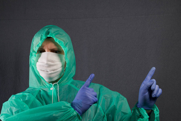 A man in a protective gauze mask and a raincoat with a hood. In rubber gloves. Holds hands in front of him, with index fingers shows up, over the shoulder. Simple protection against viral infection. It is on a gray background. - Foto, Imagem