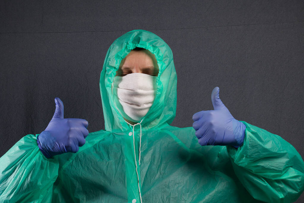A man in a protective gauze mask and a raincoat with a hood. In rubber gloves. Holds hands in front of him, thumbs up. Simple protection against viral infection. It is on a gray background. - Photo, image