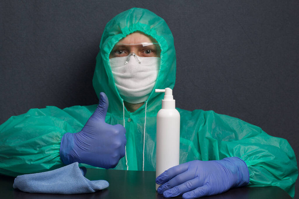 A man in a protective gauze mask, glasses and a raincoat with a hood. Hands in rubber gloves. Sits at the table, shows thumb up. Before him is a spray gun and a rag. Symbol of the global epidemic. It is on a gray background. - Фото, зображення