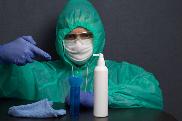 A man in a protective gauze mask, glasses and a raincoat with a hood. Hands in rubber gloves. Sits at a table, points a finger at a spray, a brush and a rag. Symbol of the global epidemic. It is on a gray background. - Photo, image