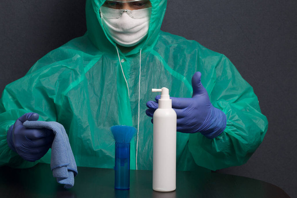 A man in a protective gauze mask, glasses and a raincoat with a hood. Hands in rubber gloves. He sits at a table, in front of him is a spray bottle, a brush and a rag. Symbol of the global epidemic. It is on a gray background. - Foto, immagini