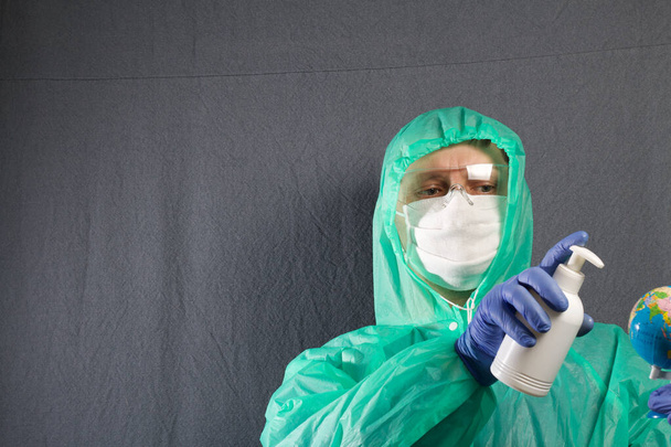 A man in a protective gauze mask, glasses and a raincoat with a hood. He holds the globe in his hand and processes it from the atomizer. Hands in rubber gloves. Symbol of the global epidemic. It is on a gray background. - Photo, Image