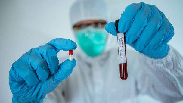 Γιατρός στο εργαστήριο κρατώντας ένα χάπι και σωλήνα εξέτασης αίματος σε λευκό φόντο. Το Covid-19 προσπαθεί να βρει ένα φάρμακο κατά του ιού για τη θεραπεία των μολυσμένων. Εμβόλιο για τη θεραπεία της έννοιας coronavirus. - Φωτογραφία, εικόνα