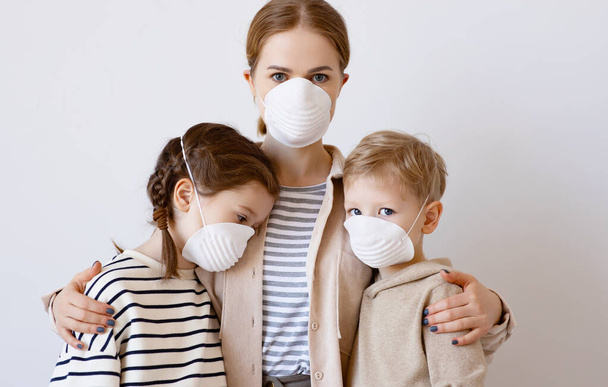 Adult woman in medical mask hugging and protecting sad girl and boy from contagious coronavirus disease against gray wal - Photo, Image