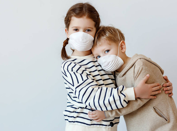 Kleines Mädchen umarmt und stützt Bruder in medizinischer Maske, während es während der Coronavirus-Pandemie vor grauer Wand steht - Foto, Bild