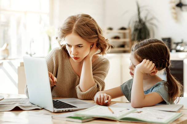 Сконцентрированная молодая женщина в повседневной одежде читает данные на ноутбуке, сидя с дочерью за столом с учебниками и тетрадью и изучает онлин
 - Фото, изображение