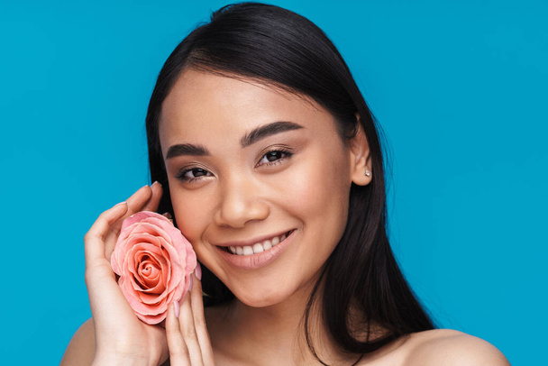 Photo of a pretty happy cheerful asian young woman posing isolated over blue wall background with flower. - Foto, Imagem