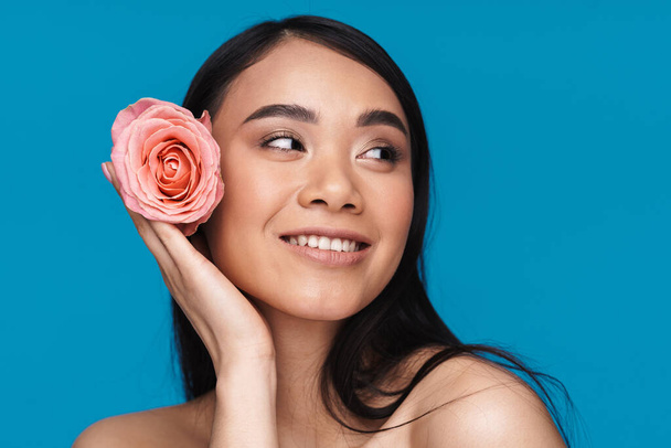 Photo of a pretty happy cheerful asian young woman posing isolated over blue wall background with flower. - Photo, Image