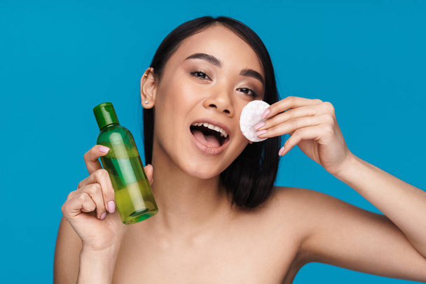 Photo of asian positive young woman posing isolated over blue wall background take care of her skin with cleansing water and cotton pad. - Foto, afbeelding