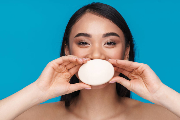 Photo of asian positive happy young woman posing isolated over blue wall background holding soap covering mouth. - Foto, afbeelding