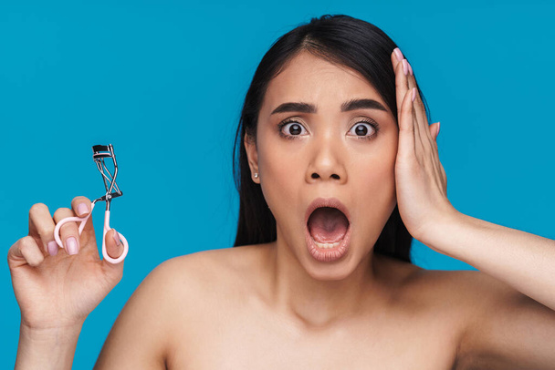 Photo of asian shocked surprised young woman posing isolated over blue wall background holding eyelash curler. - Fotografie, Obrázek