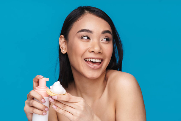 Photo of asian positive young woman posing isolated over blue wall background holding mousse cleansing foam. - Photo, Image