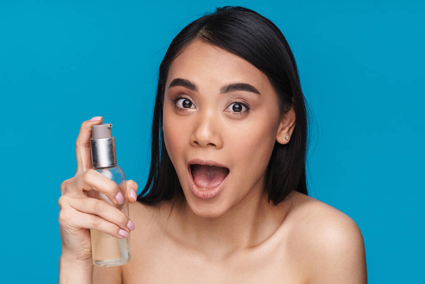 Photo of asian emotional young woman posing isolated over blue wall background holding bottle with cosmetic oil. - Zdjęcie, obraz