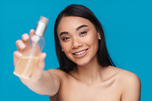 Photo of asian cheerful young woman posing isolated over blue wall background showing bottle with cosmetic oil. - 写真・画像