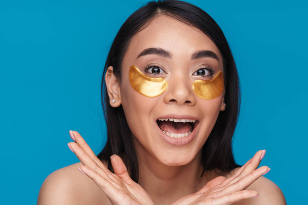 Photo of asian smiling optimistic young woman posing isolated over blue wall background take care of undereye skin with patches. - Фото, зображення