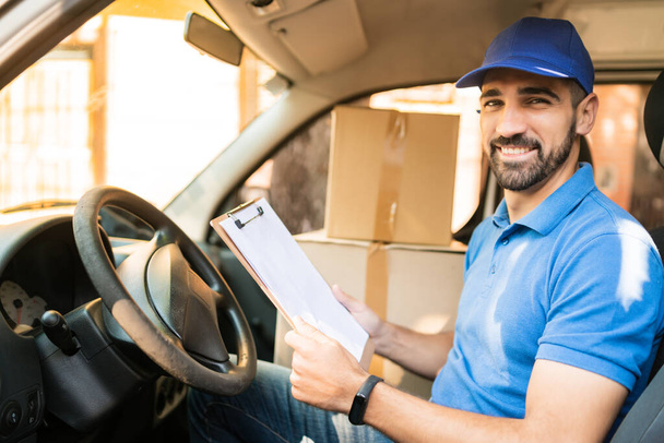 Portrait of a delivery man checking the delivery list while sitting in van. Delivery and shipping concept. - Foto, Imagen