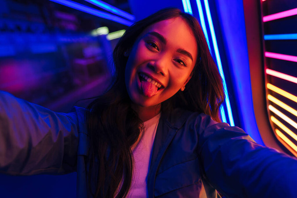 Photo of young optimistic woman posing over neon lights take a selfie by camera showing tongue. - Fotografie, Obrázek