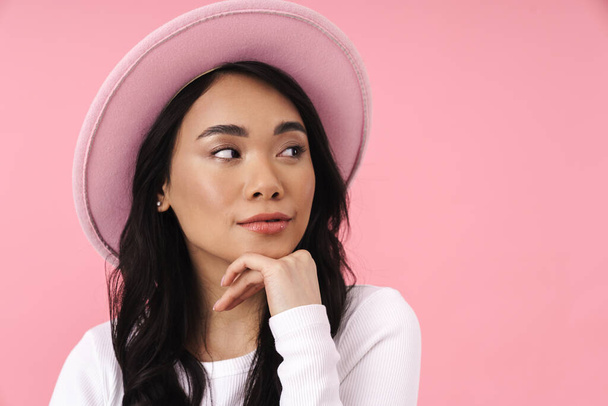Attractive smiling young asian woman wearing casual clothes standing isolated over pink background, posing in hat - Zdjęcie, obraz