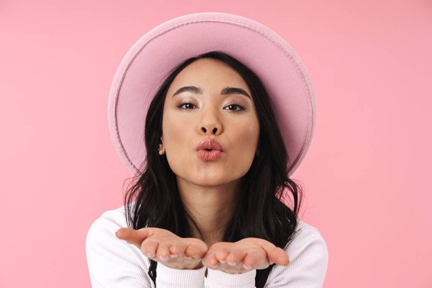 Image of young brunette asian woman wearing hat blowing air kiss at camera isolated over pink background - Zdjęcie, obraz