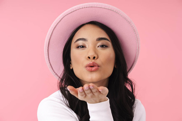 Image of young brunette asian woman wearing hat blowing air kiss at camera isolated over pink background - Fotoğraf, Görsel