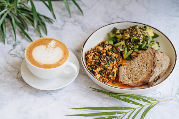 Tofu scramble with pumpkin seeds, avocado salad with cucumber and basilic served with rye toasts in white plate on a marble background - Фото, изображение